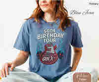 Woman wearing a blue Comfort Colors t-shirt with a “50th Birthday Tour Gen X” graphic featuring a red electric guitar, lightning bolts, and vintage-style text, seated cross-legged in a relaxed pose.