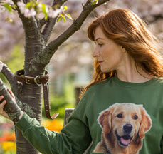 A woman sits on a bench in a peaceful spring garden, quietly reflecting as she gazes at a blooming tree. Wrapped around a small branch is her deceased dog’s collar, symbolizing a personal pet memorial. Her green sweatshirt features a smiling portrait of her beloved golden retriever, capturing the emotional bond and deep grief of losing a furry friend. The scene conveys themes of coping with pet loss, honoring a pet’s memory, healing after pet death, and the lasting love we carry for our animals.