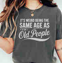 Woman wearing vintage-style Comfort Colors t-shirt with bold white text that reads “It’s Weird Being the Same Age as Old People,” in relaxed, casual pose with long wavy hair.