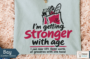 	Bay green shirt with a grocery bag graphic reading “I’m getting stronger with age. I can now lift $200 worth of groceries with one hand.”