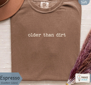 	Espresso brown aging humor shirt with small white lettering reading “older than dirt” across the chest.